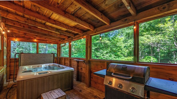 Eine Holzblockhütte mit Whirlpool, Grill und großen Fenstern mit Blick auf einen Wald.