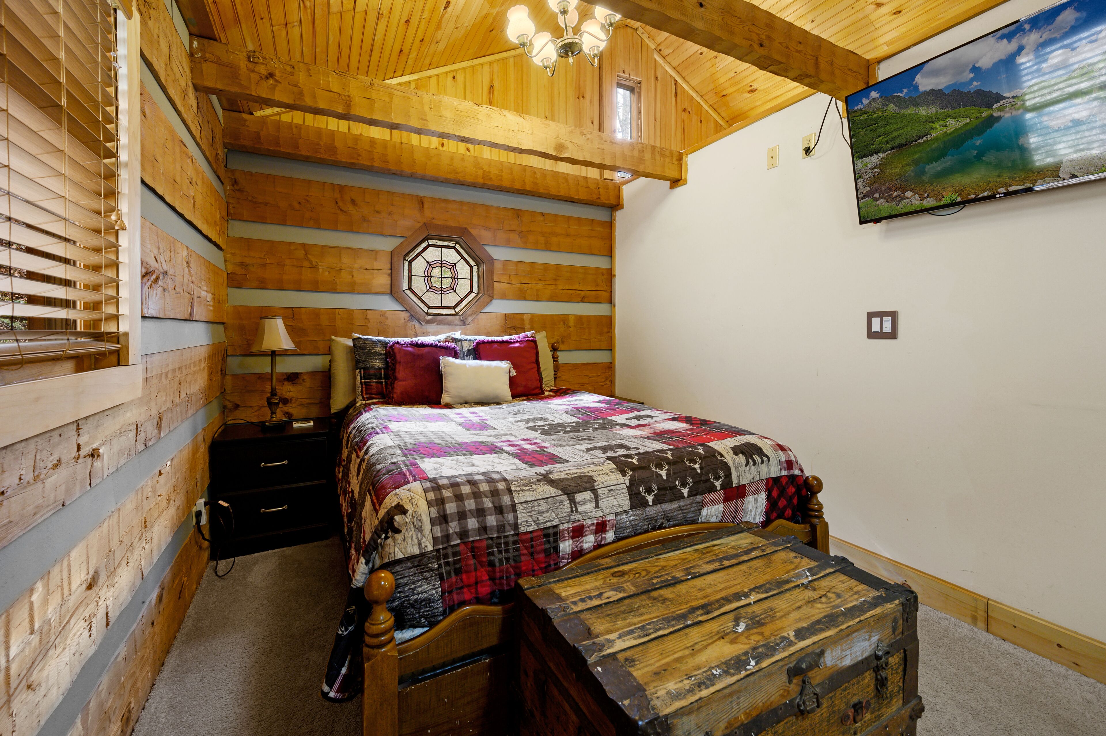 A cozy bedroom with a wooden ceiling, a bed with a quilt, a nightstand, a lamp, and a wooden chest.