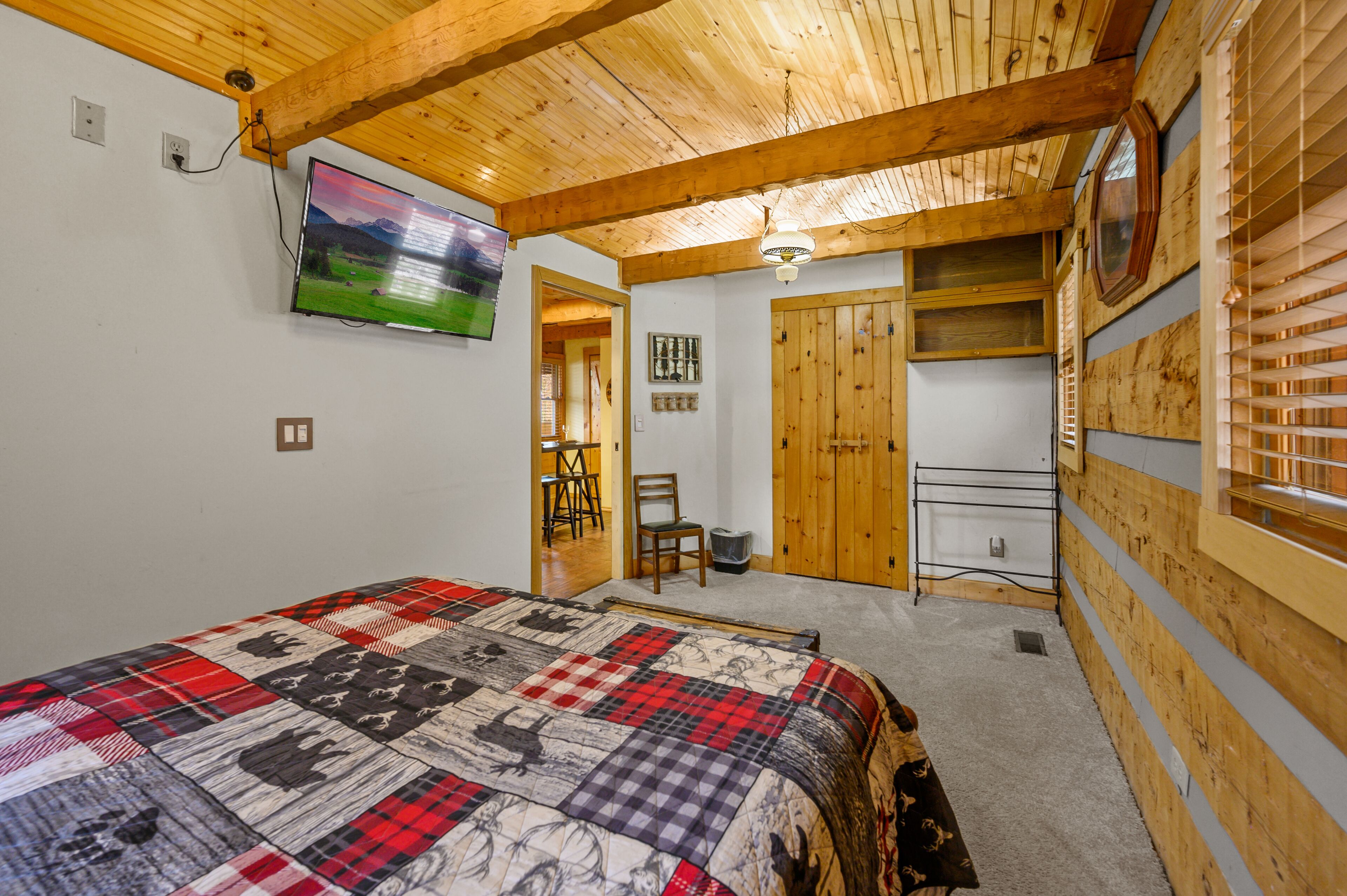 A bedroom with a wooden ceiling, a bed with a plaid quilt, a TV on the wall, and a wooden door.