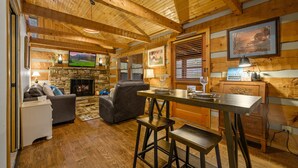 Ein gemütliches Blockhaus-Interieur mit Kamin aus Stein, Holzbalkendecke, Fernseher und einer Bar.