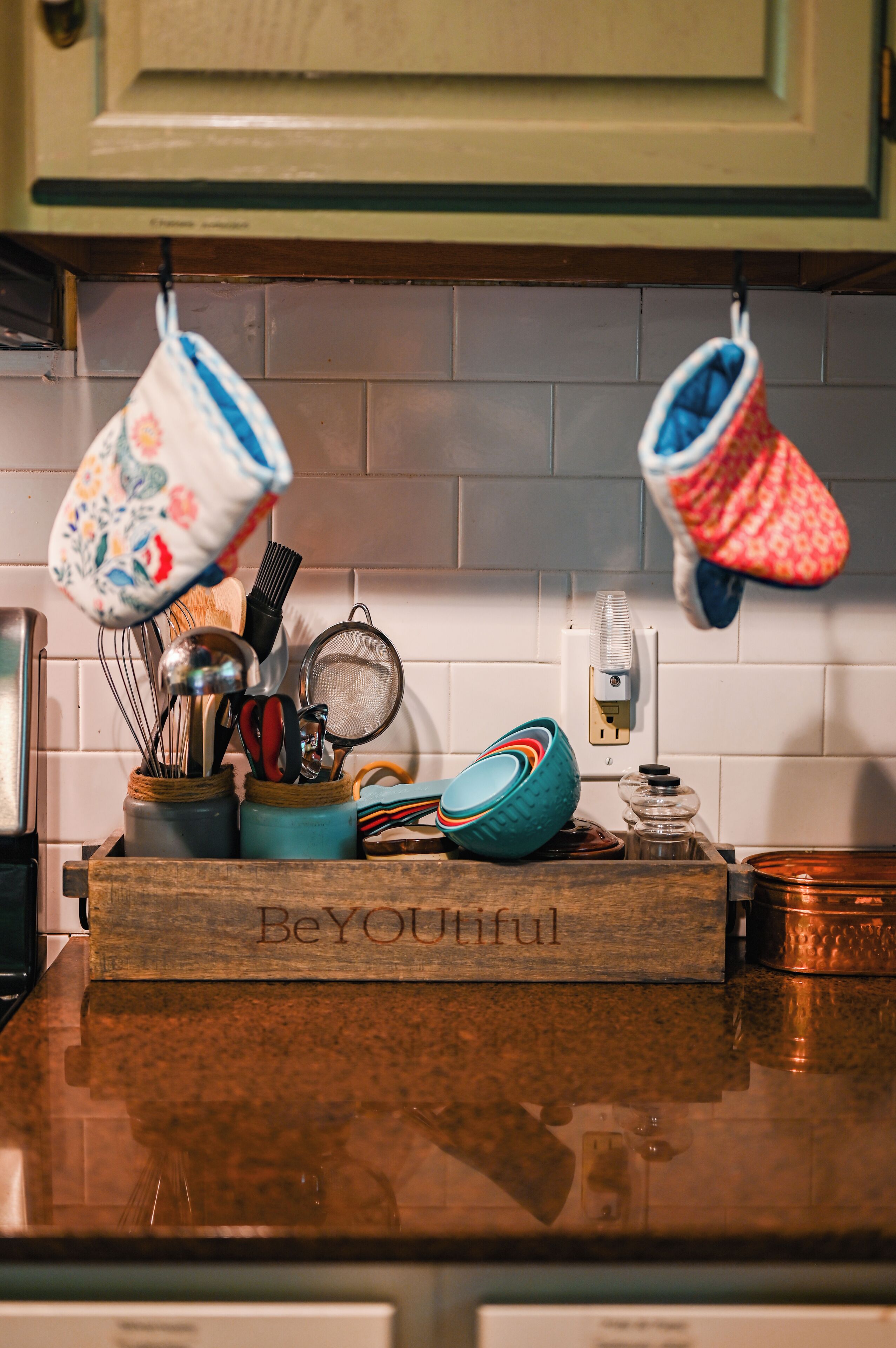 Una encimera de cocina con una tabla de cortar de madera que dice 'BeYOUtiful', un especiero y utensilios de cocina colgando.