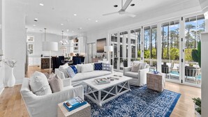 A spacious living room with a large white sofa, a coffee table, and a blue patterned rug. There is a kitchen area in the background with white cabinets and a dining table.