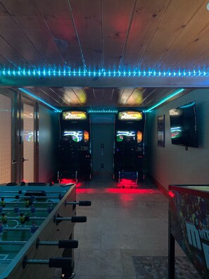 Une pièce équipée de deux bornes d’arcade, d’un baby-foot et d’un éclairage à bandes LED.
