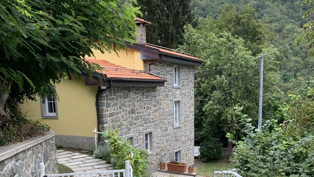 Una casa de piedra con techo de tejas rojas, cerca blanca y jardín con plantas en macetas.