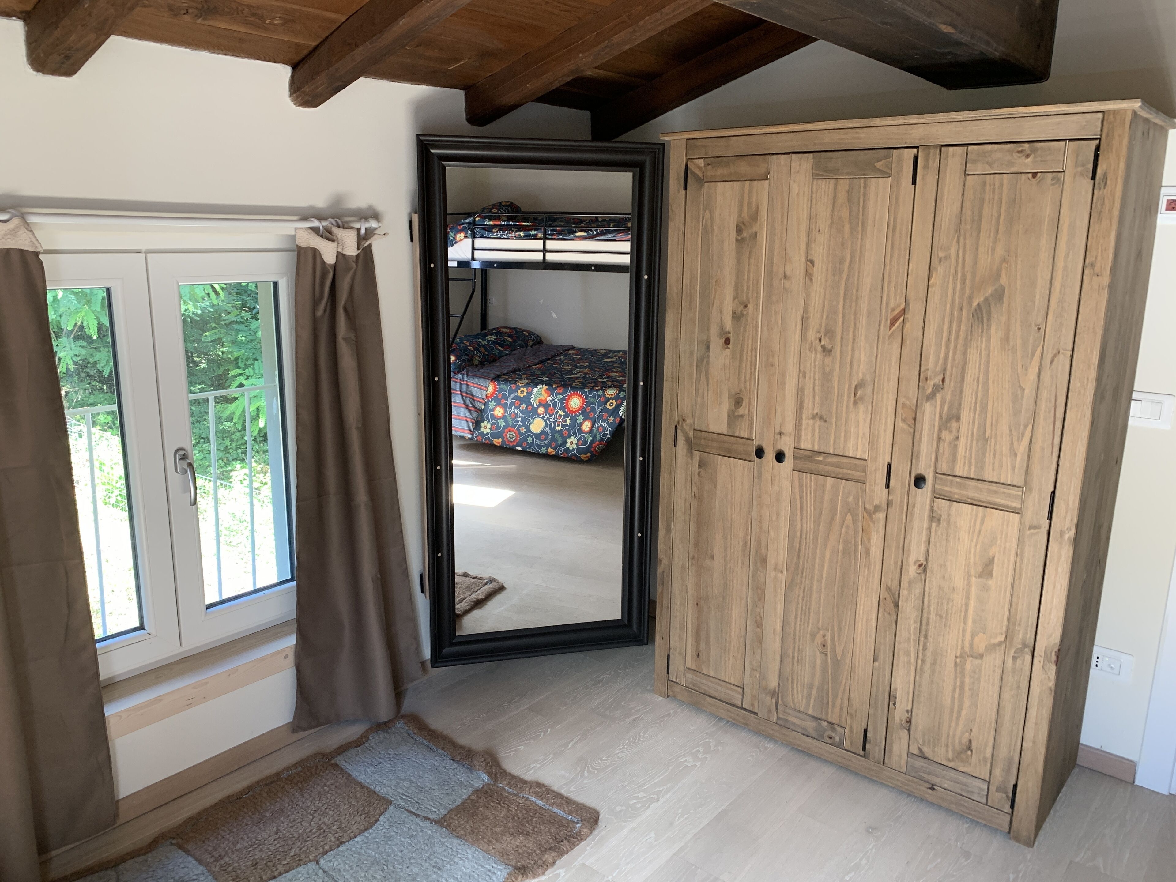 A room with a wooden wardrobe, a bed with a floral bedspread, and a window with curtains.