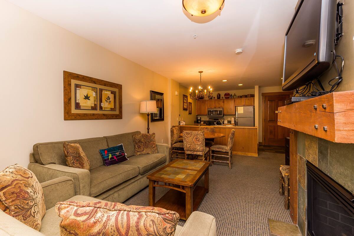 A living room with a sofa, a coffee table, and patterned cushions. There is a fireplace and a television mounted on the wall.