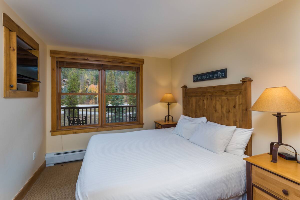 A bedroom with a large bed, a wooden headboard, a window with a view of trees, and a small table with a lamp.