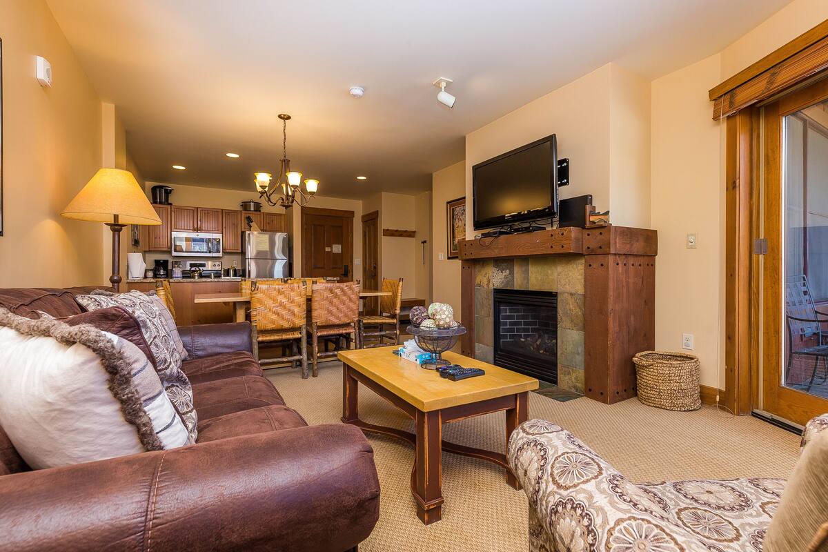 A living room with a sofa, a wooden coffee table, and a fireplace. There is a kitchen in the background with a microwave and a dining area with chairs.