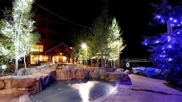 Zona de hidromasaje con piscina, rodeada de rocas y árboles, y un edificio con luces al fondo.