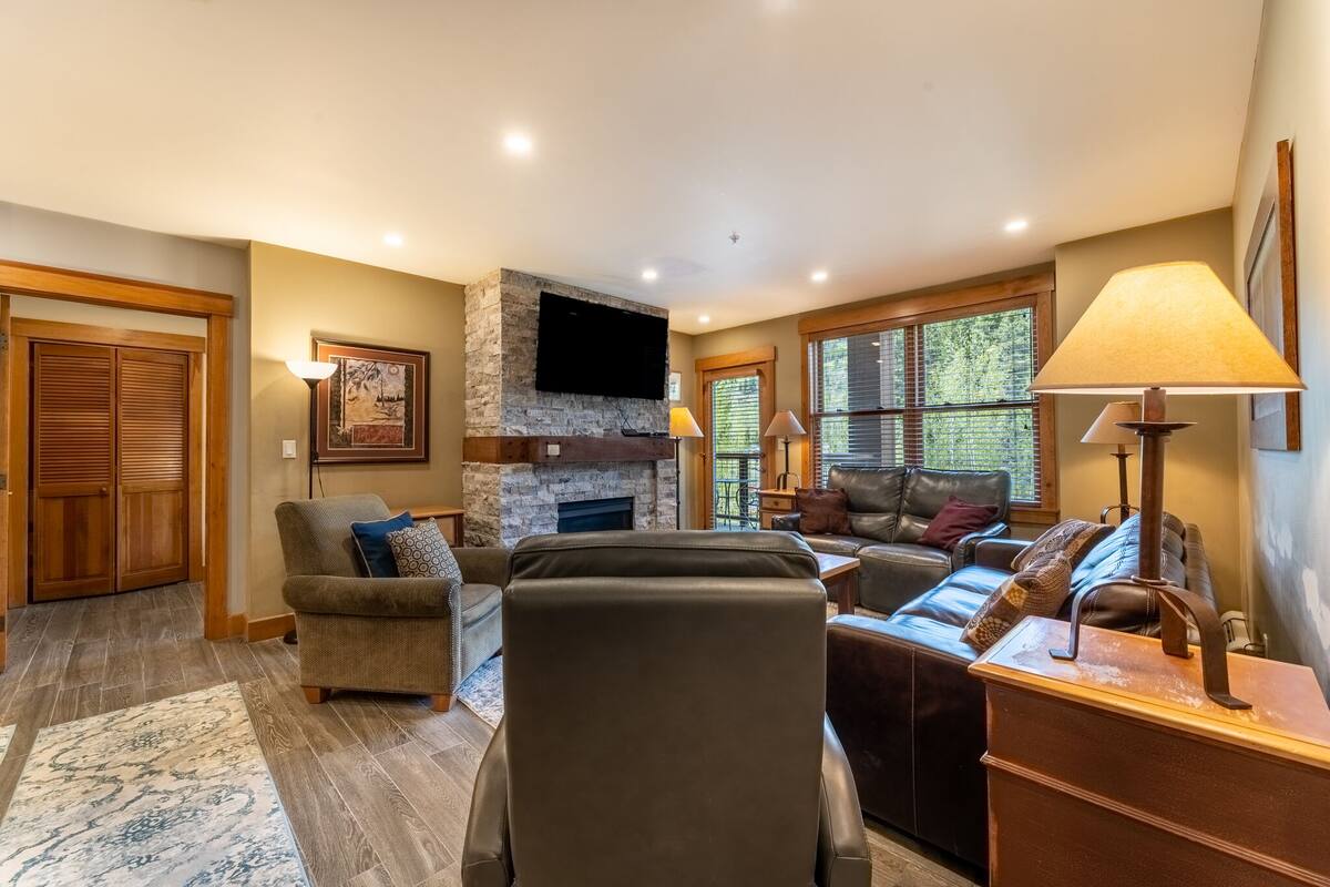 A spacious living room with a stone fireplace, leather sofas, and a flat-screen TV mounted above it.