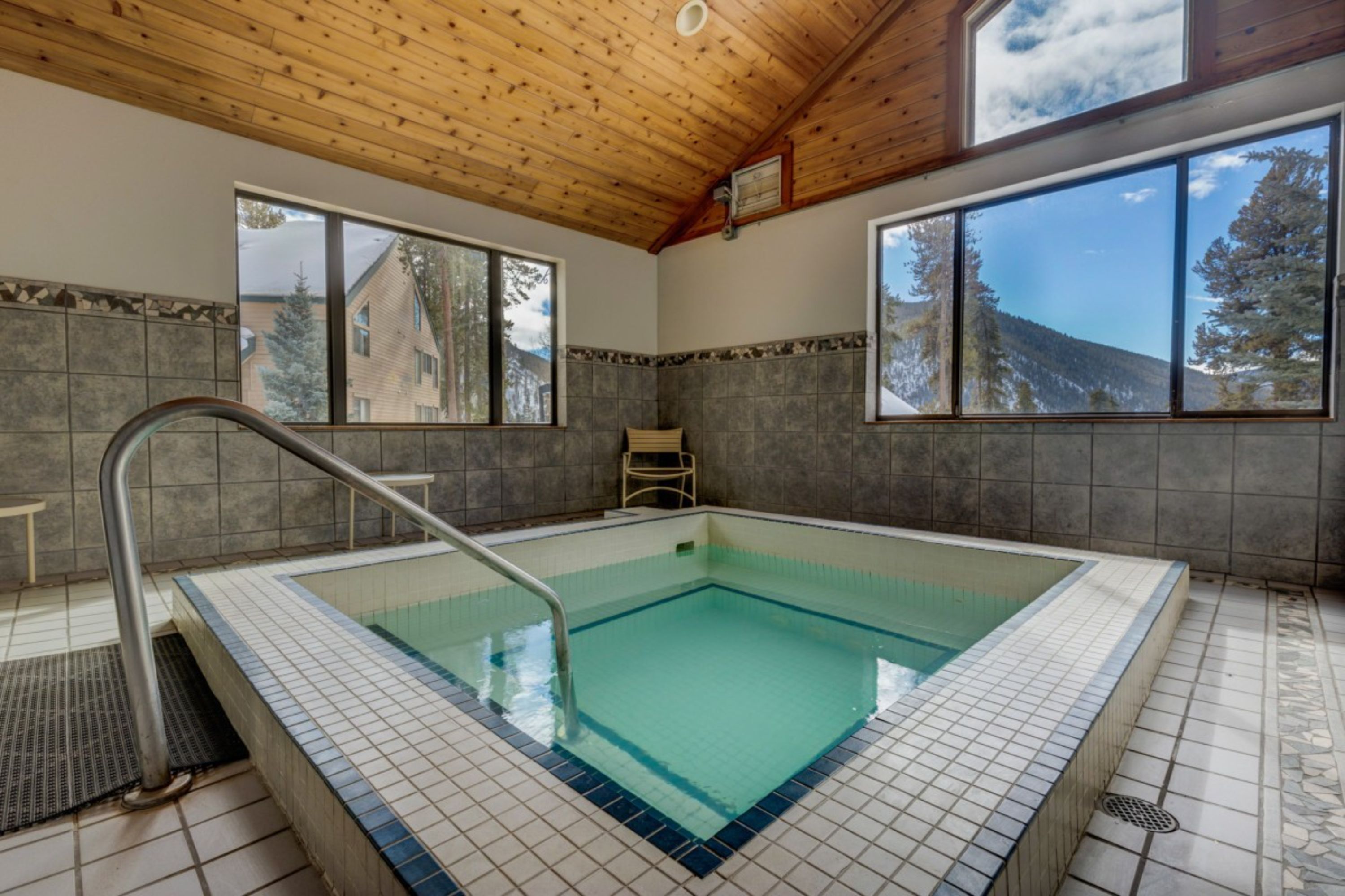 A swimming pool with tiled walls and a wooden ceiling, large windows, and a view of a building and trees.