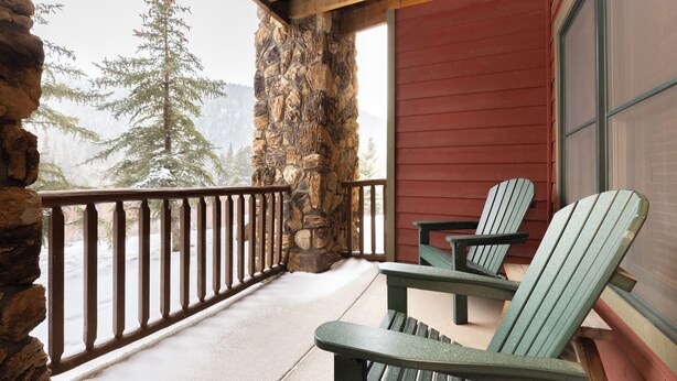 A stone wall, wooden deck, and two Adirondack chairs on a porch with a view of snow-covered trees.