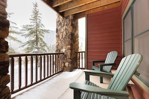 A stone wall, wooden deck, and two Adirondack chairs on a porch with a view of snow-covered trees. - Hidden River Lodge #5941 by Summit County Mountain Retreats (Keystone)