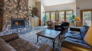 A spacious living area with a stone fireplace, leather sofas, a coffee table, and large windows offering a view of the outdoors.