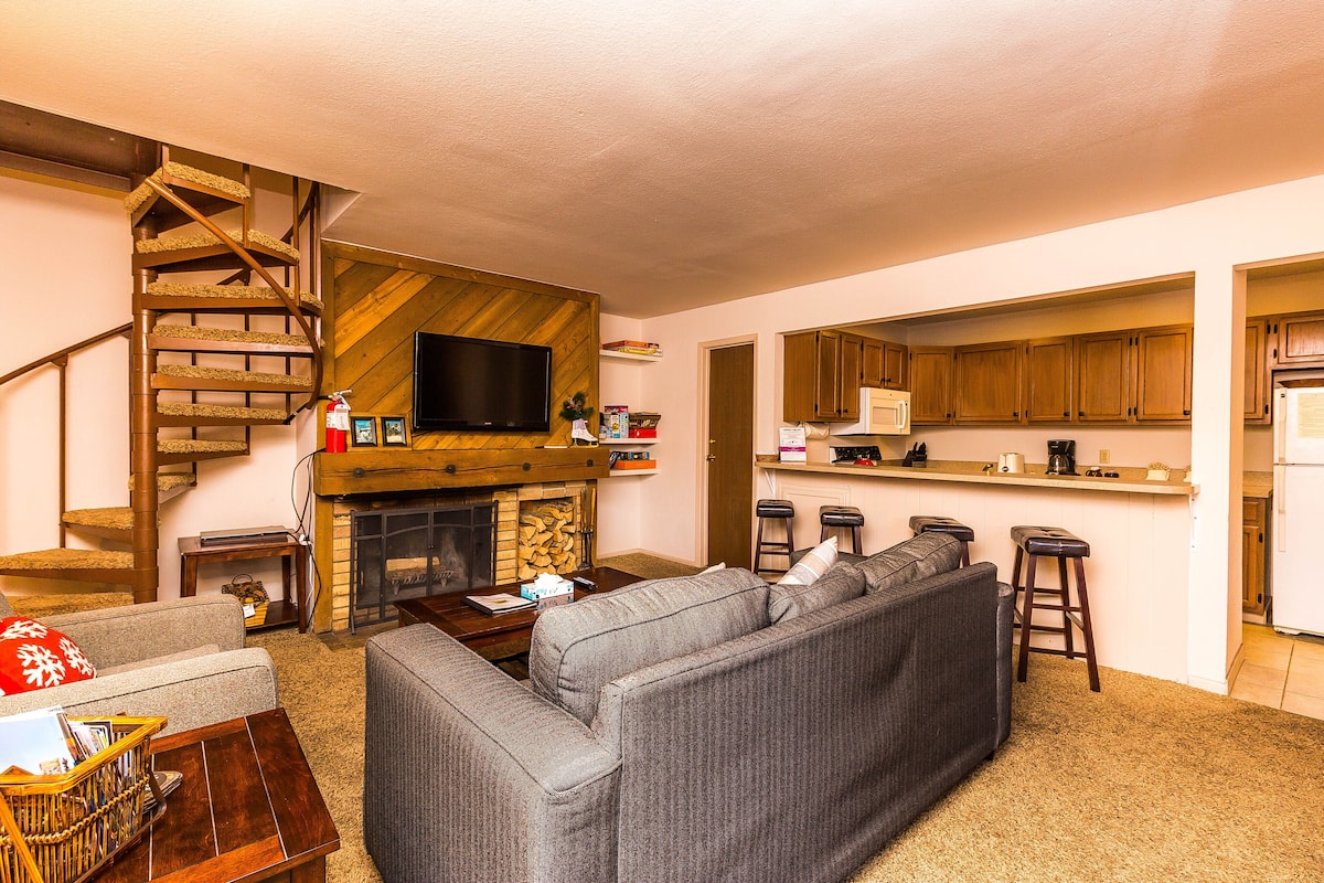 A living room with a fireplace, a sofa, a coffee table, and a TV. There is a kitchen area with a microwave and a refrigerator.
