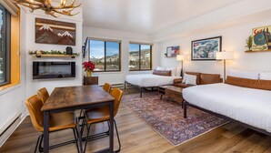 Een moderne hotelkamer met een houten eettafel, twee eenpersoonsbedden, een bank en een groot raam met uitzicht op de bergen.