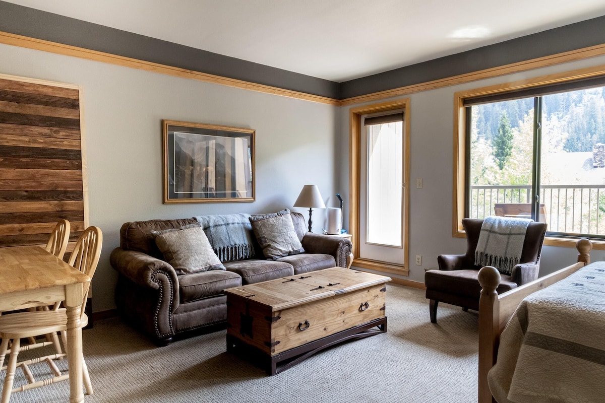 A living room with a wooden coffee table, a brown leather sofa, a dining table with chairs, and a large window with a view of trees.