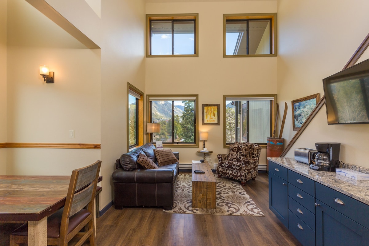 A spacious living area with a dining table, leather sofa, patterned armchair, and a kitchen area with a coffee maker.