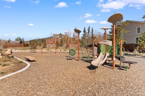 A playground with various play structures, including a slide and swings, surrounded by a gravel area with trees and a building in the background. - Dillon Valley East by Summit County Mountain Retreats (Dillon)
