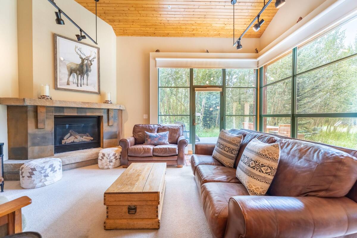 A cozy living room with a fireplace, leather sofas, and a large window offering a view of the outdoors.
