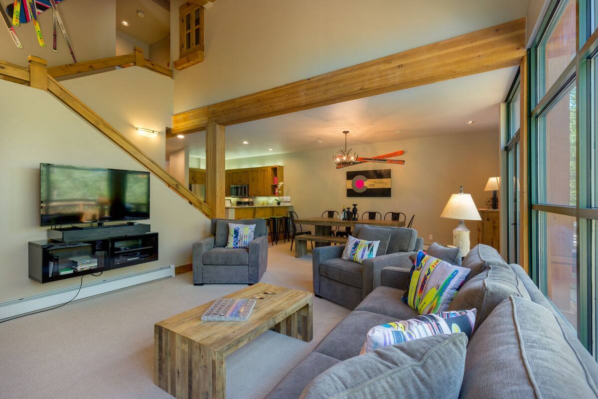 A spacious living room with a large gray sofa, two armchairs, a wooden coffee table, and a television on a black stand. A staircase with wooden railings leads to an upper level.