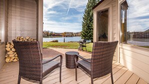 Une terrasse couverte, meublée avec des meubles en osier, dotée d’un barbecue et offrant une vue sur un lac et des bâtiments.