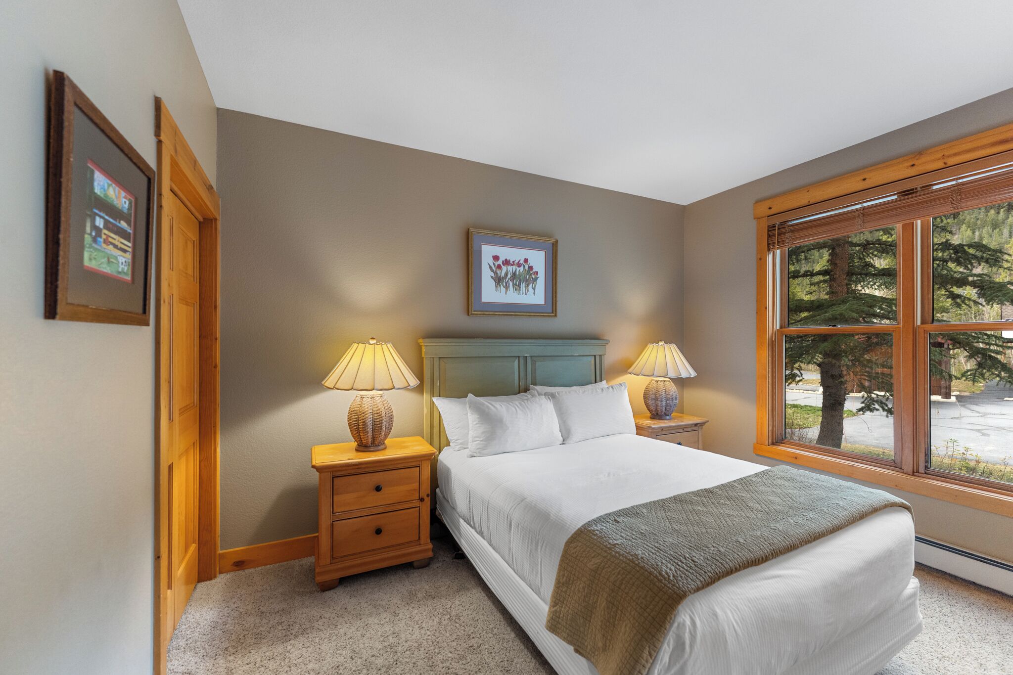 A bedroom with a bed, two bedside tables with lamps, a framed picture on the wall, and a window with a view of trees.