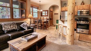 A spacious living room with a leather sofa, wooden coffee table, and a dining area with wooden chairs and a chandelier. The kitchen features stainless steel appliances and wooden cabinets. - Trapper's Crossing #8770 by Summit County Mountain Retreats (Keystone)
