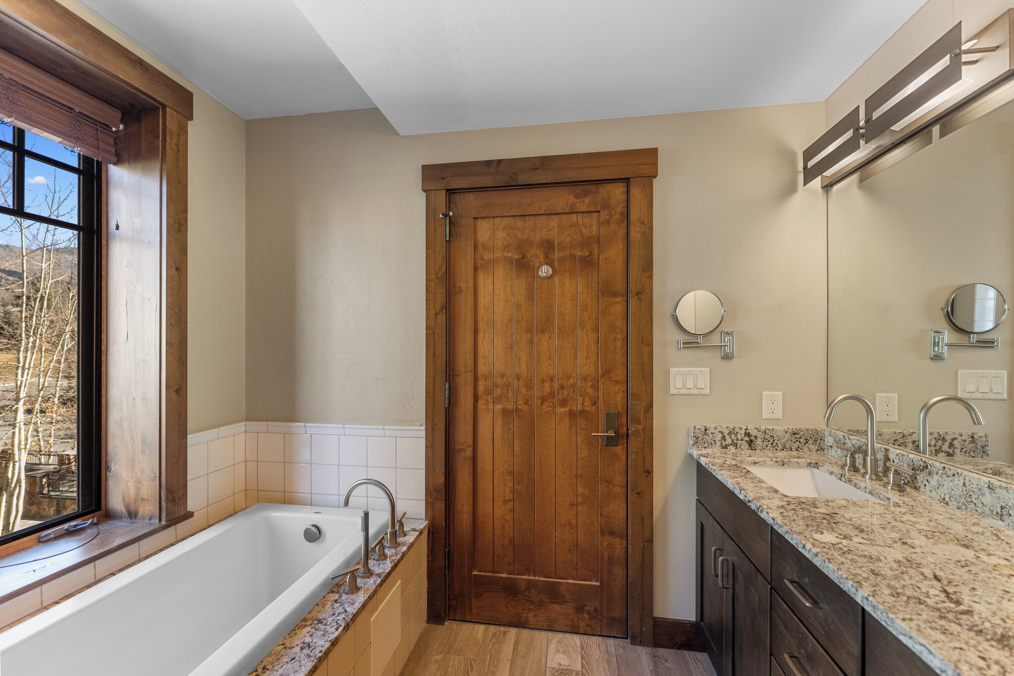 A bathroom with a wooden door, a bathtub, a sink with a granite countertop, and a window with a view of trees.