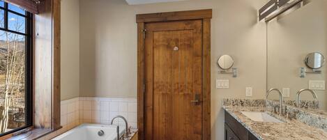 A bathroom with a wooden door, a bathtub, a sink with a granite countertop, and a window with a view of trees.