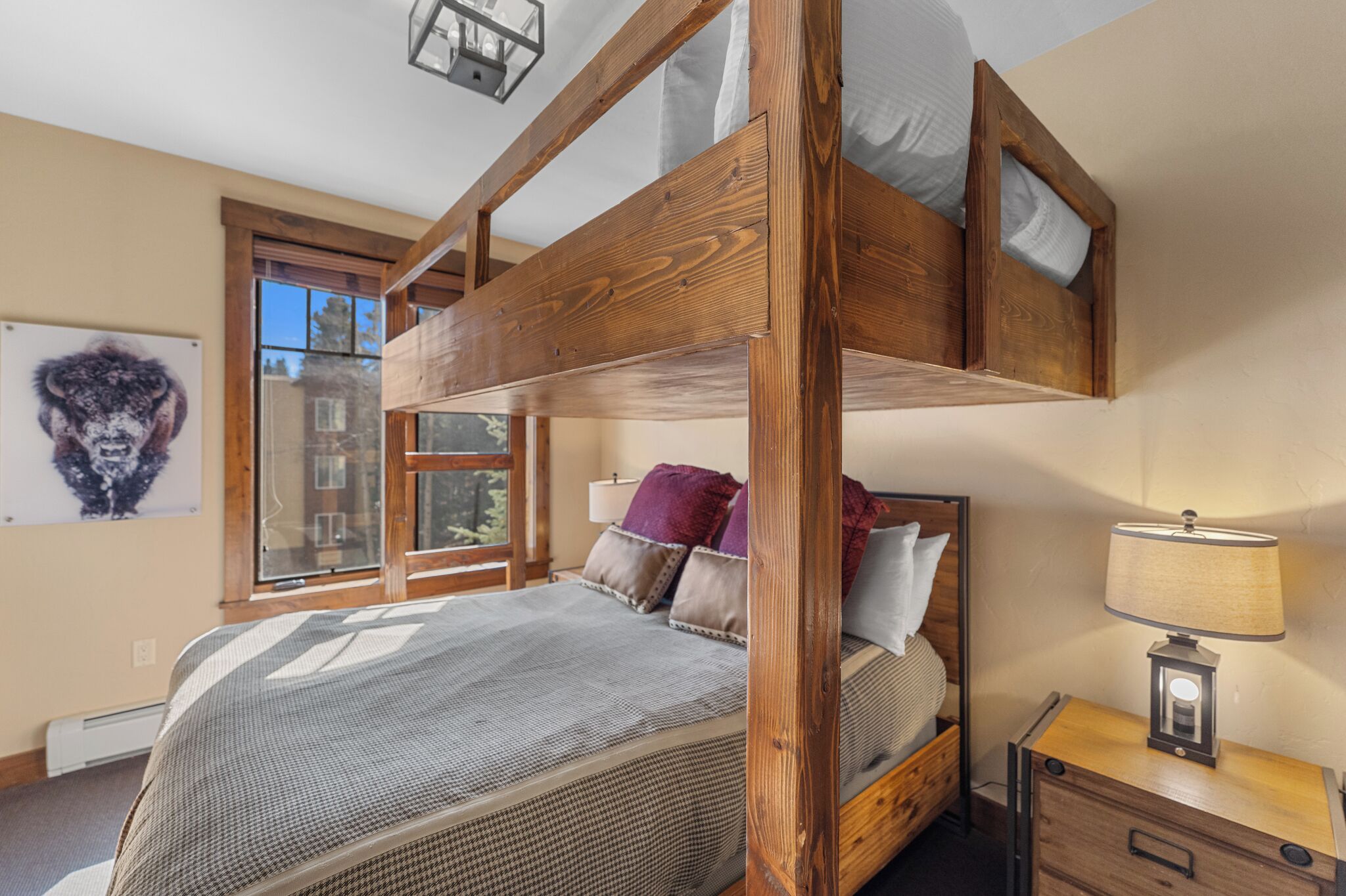 A bunk bed with a wooden frame, a bedside table with a lamp, and a window with a view of buildings.
