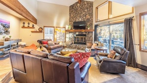 A spacious living room with leather sofas, a stone fireplace, and large windows offering a view of the outdoors. - Enclave #34 by Summit County Mountain Retreats (Keystone)