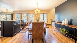A modern kitchen with a central island, wooden dining table, and chairs. There is a chandelier, large windows, and a view of trees outside. - Enclave #34 by Summit County Mountain Retreats (Keystone)