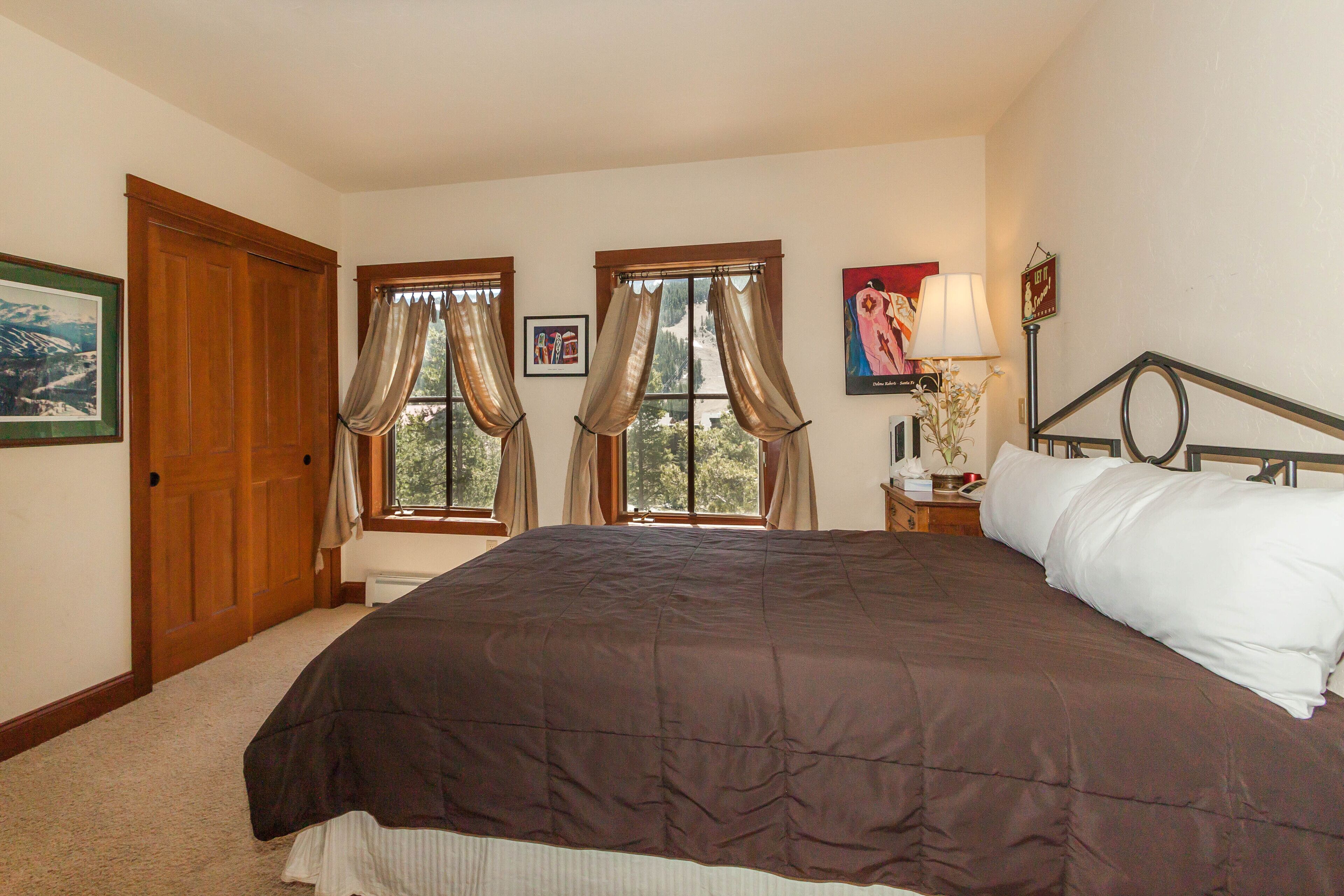 A bedroom with a large bed, two windows with curtains, a nightstand with a lamp, and a framed picture on the wall.