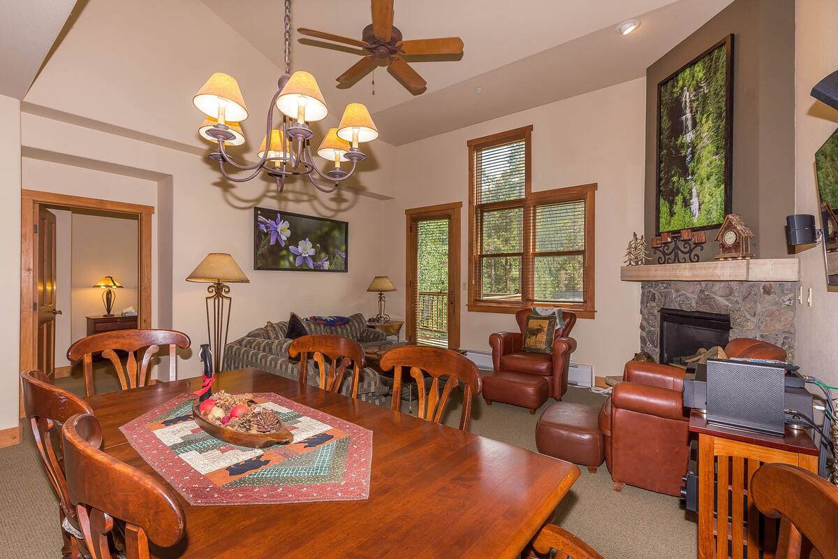 A spacious living room with a wooden dining table, comfortable seating, a fireplace, and large windows.