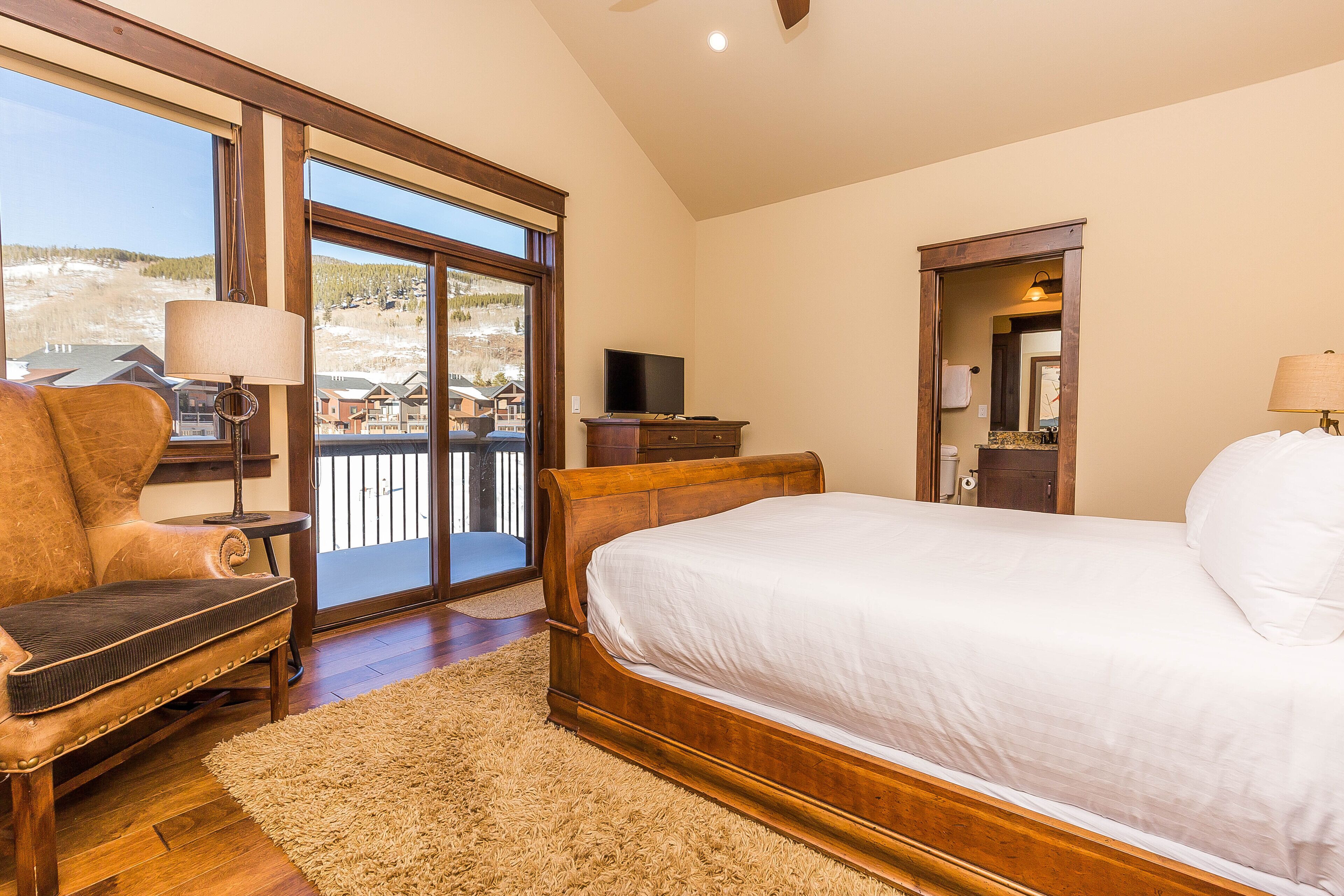 A bedroom with a large bed, a chair, a TV, and a view of a mountain landscape through a sliding door.