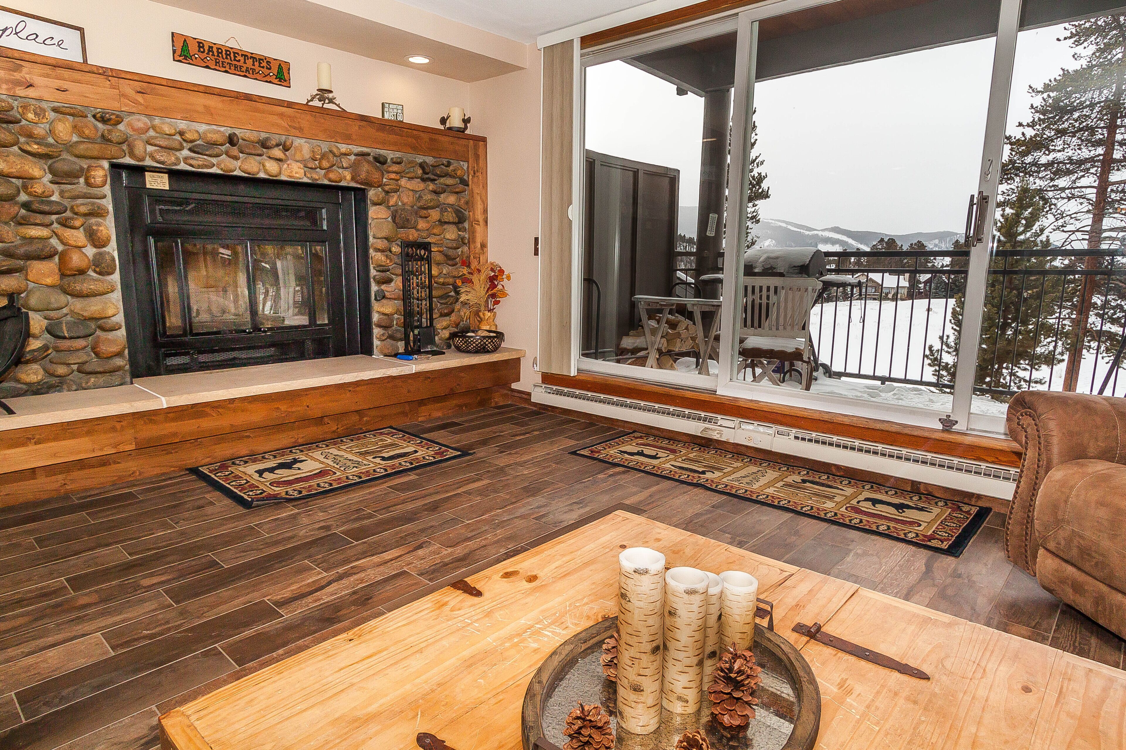 A cozy living room with a stone fireplace, wooden flooring, and a large window offering a view of the outdoors.