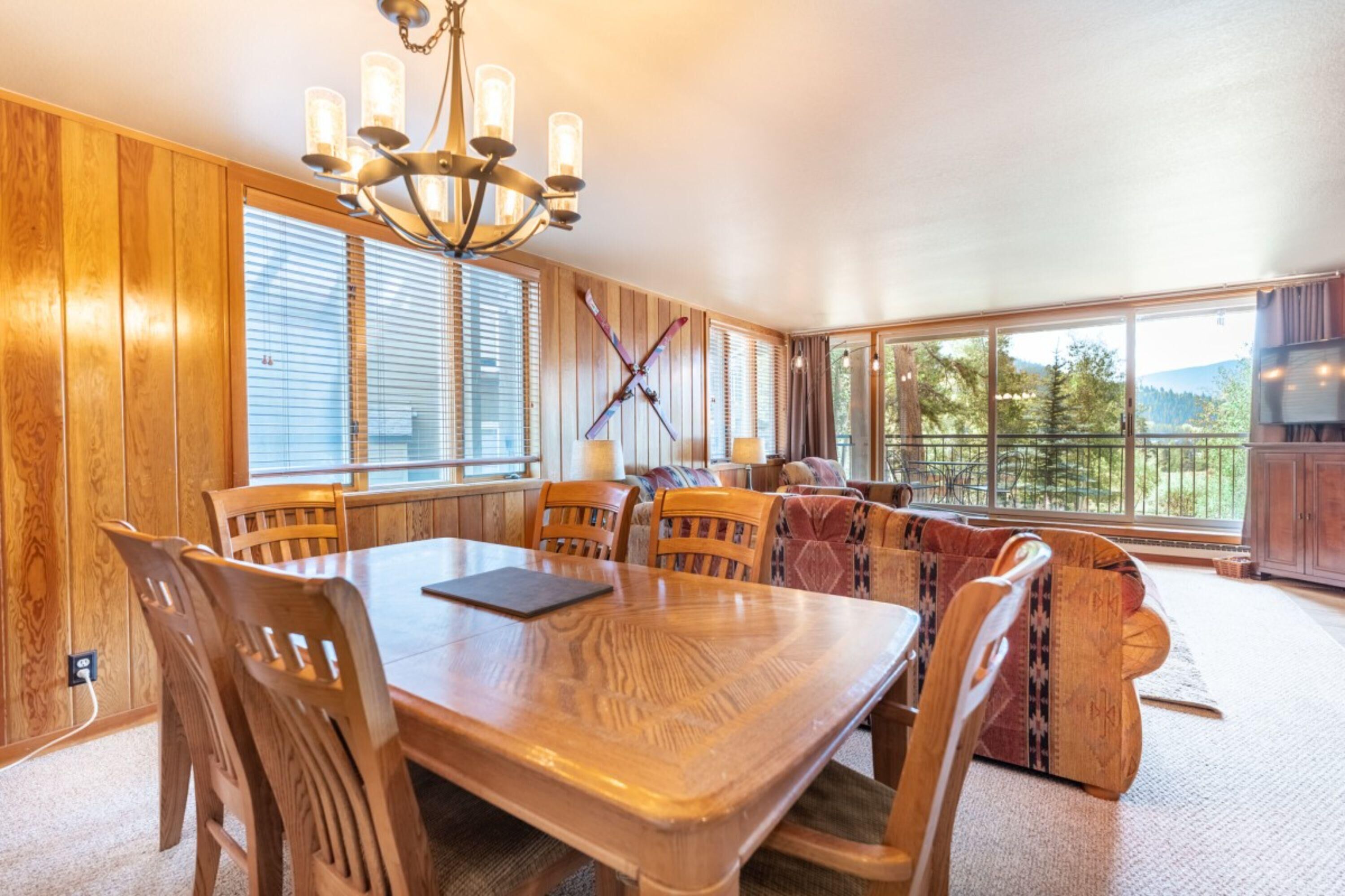 A dining area with a wooden table and chairs, a chandelier, and large windows offering a view of the outdoors.