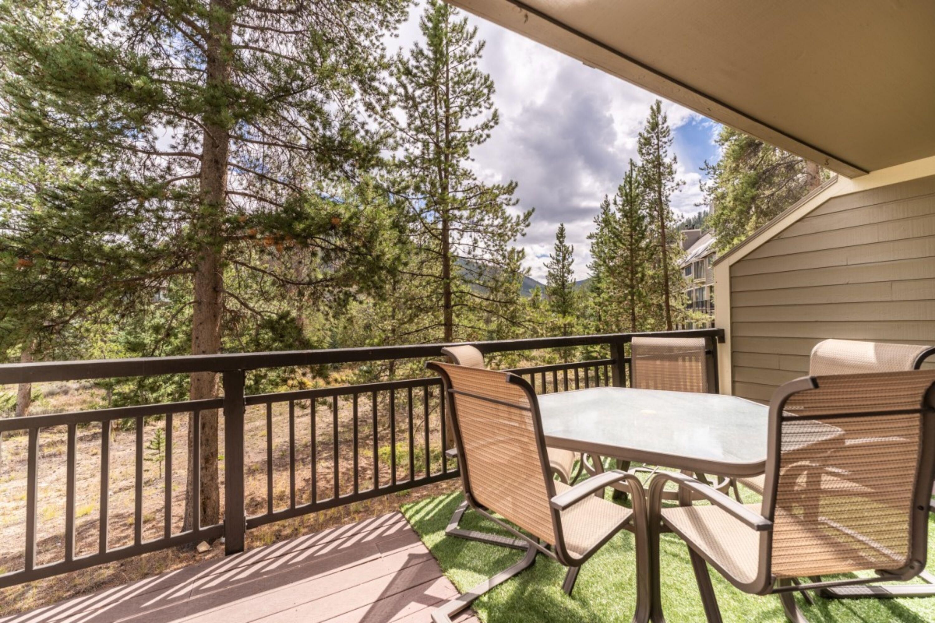A covered patio with a table and chairs, surrounded by a railing, overlooking a forested area.