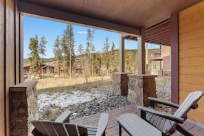 A wooden deck with Adirondack chairs, a stone fire pit, and a view of a forested area with buildings in the background.