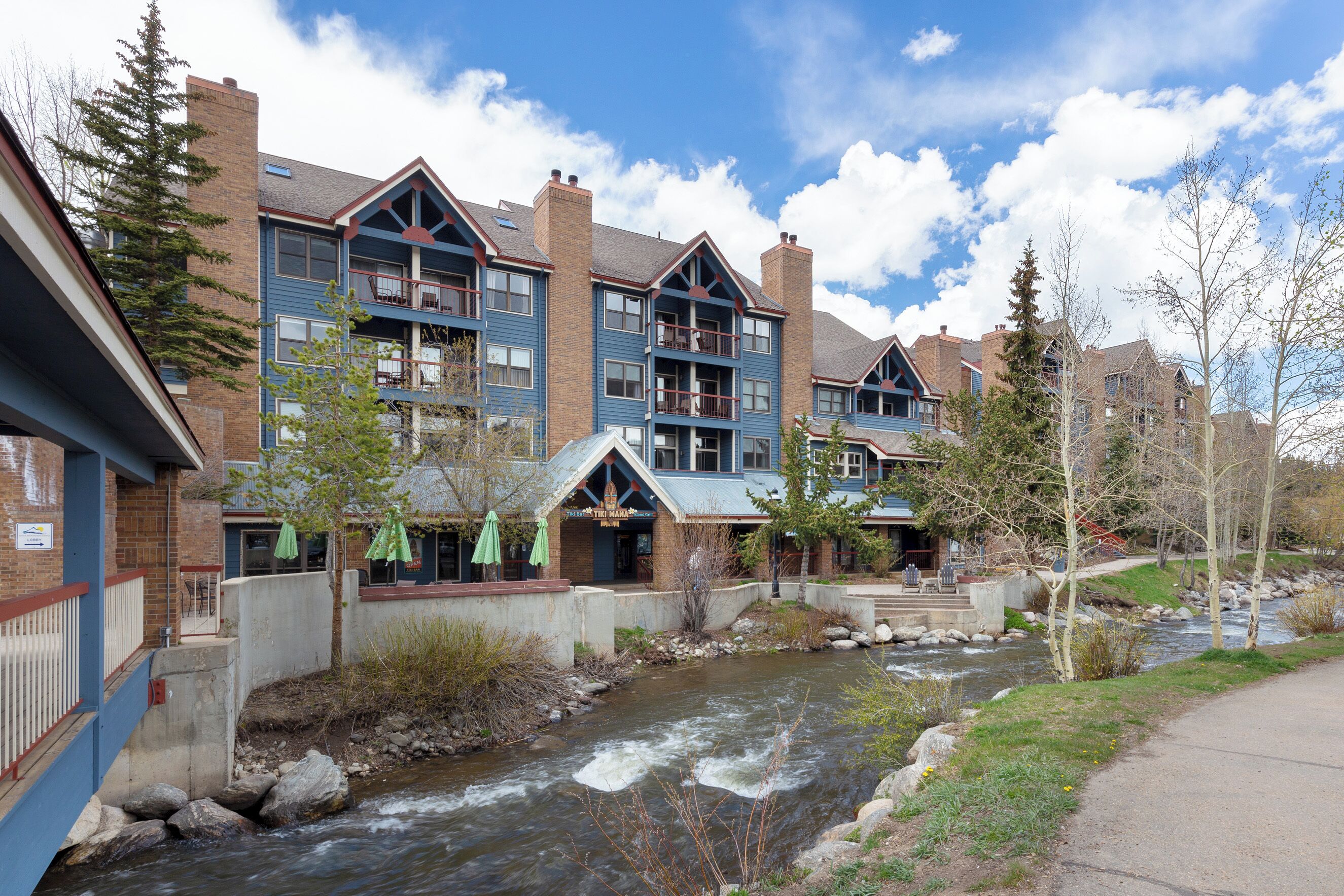 A multi-story hotel with balconies, a river in front, and outdoor seating areas.