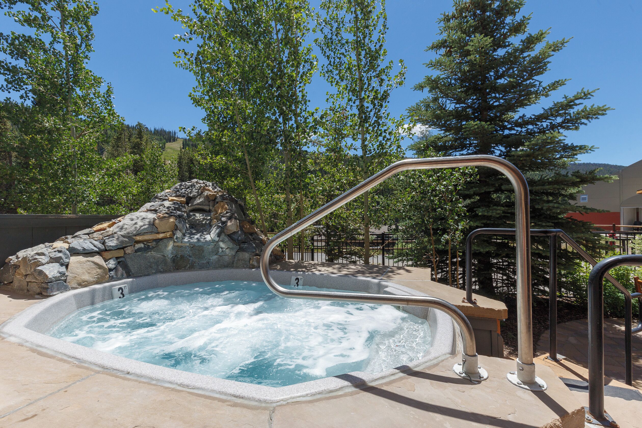 A hot tub with a stone waterfall feature, surrounded by trees and a clear sky.
