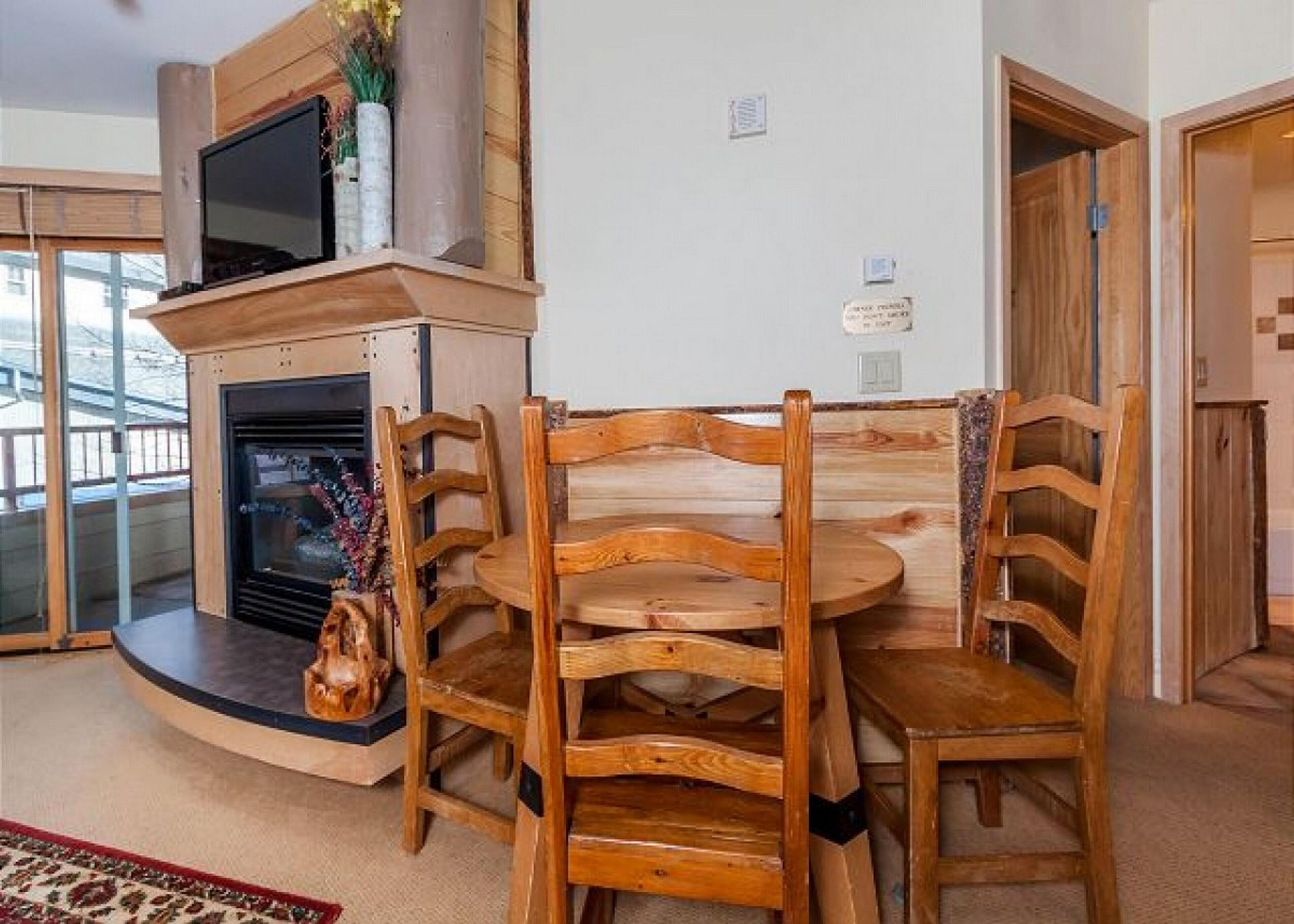 A cozy living room with a fireplace, wooden chairs, a round table, and a TV mounted on the wall.