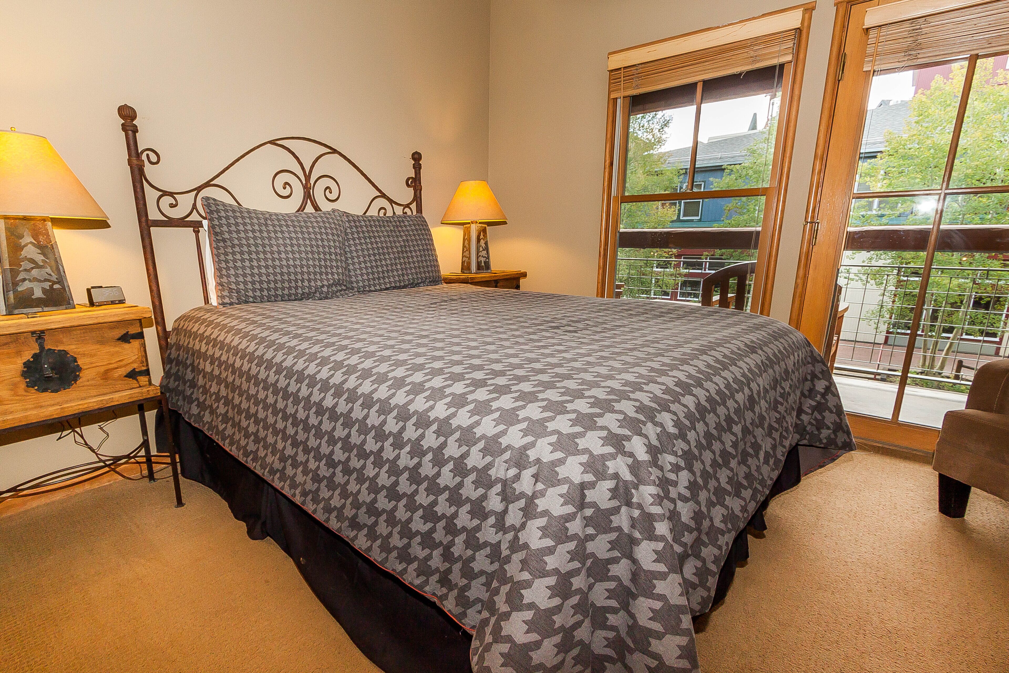 A bedroom with a bed, two bedside lamps, a nightstand, and a window with a view of greenery and buildings.