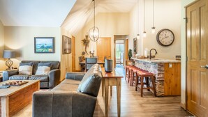 A spacious living area with a sofa, coffee table, and dining area. There is a kitchen with a stone countertop and wooden cabinets. A clock is visible on the wall. - Jackpine Lodge by Summit County Mountain Retreats (Keystone)