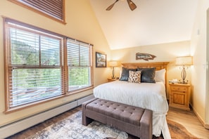 Une chambre à coucher avec un grand lit, deux tables de chevet avec des lampes, un banc et une vue sur l’extérieur grâce à de grandes fenêtres.