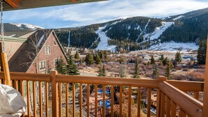 Un balcon en bois donnant sur une station de ski, avec vue sur la montagne, une piscine et un bâtiment rouge.