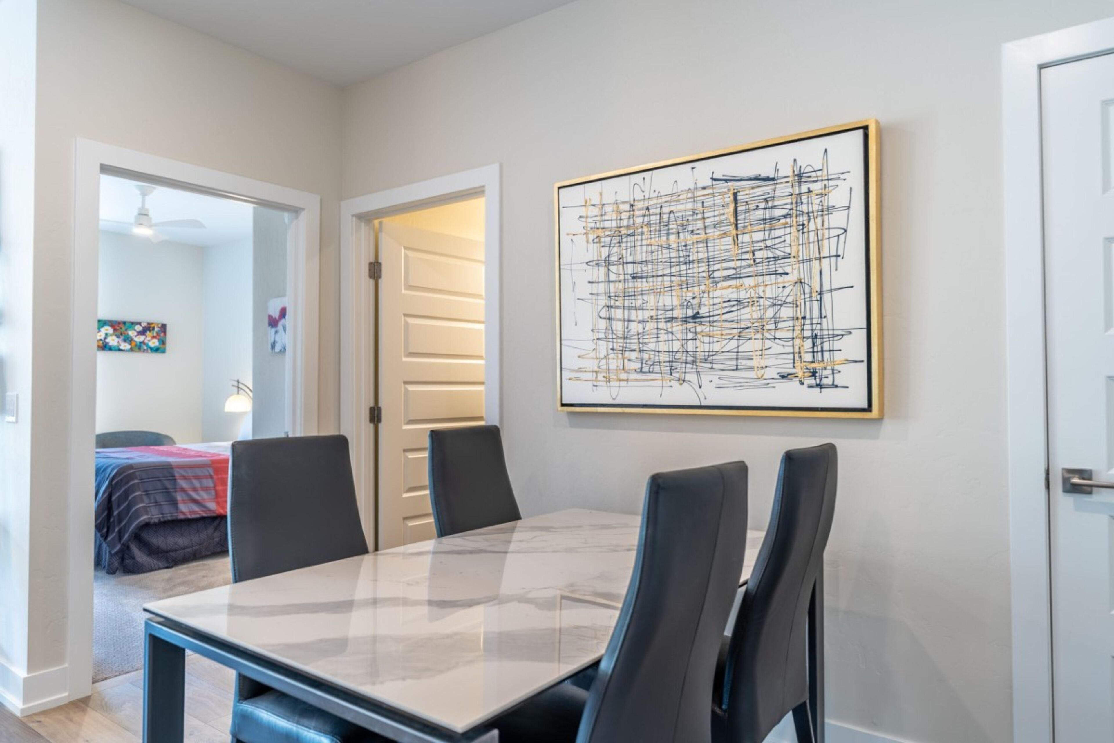 A dining area with a marble table and black chairs, a framed abstract painting on the wall, and a door leading to another room.