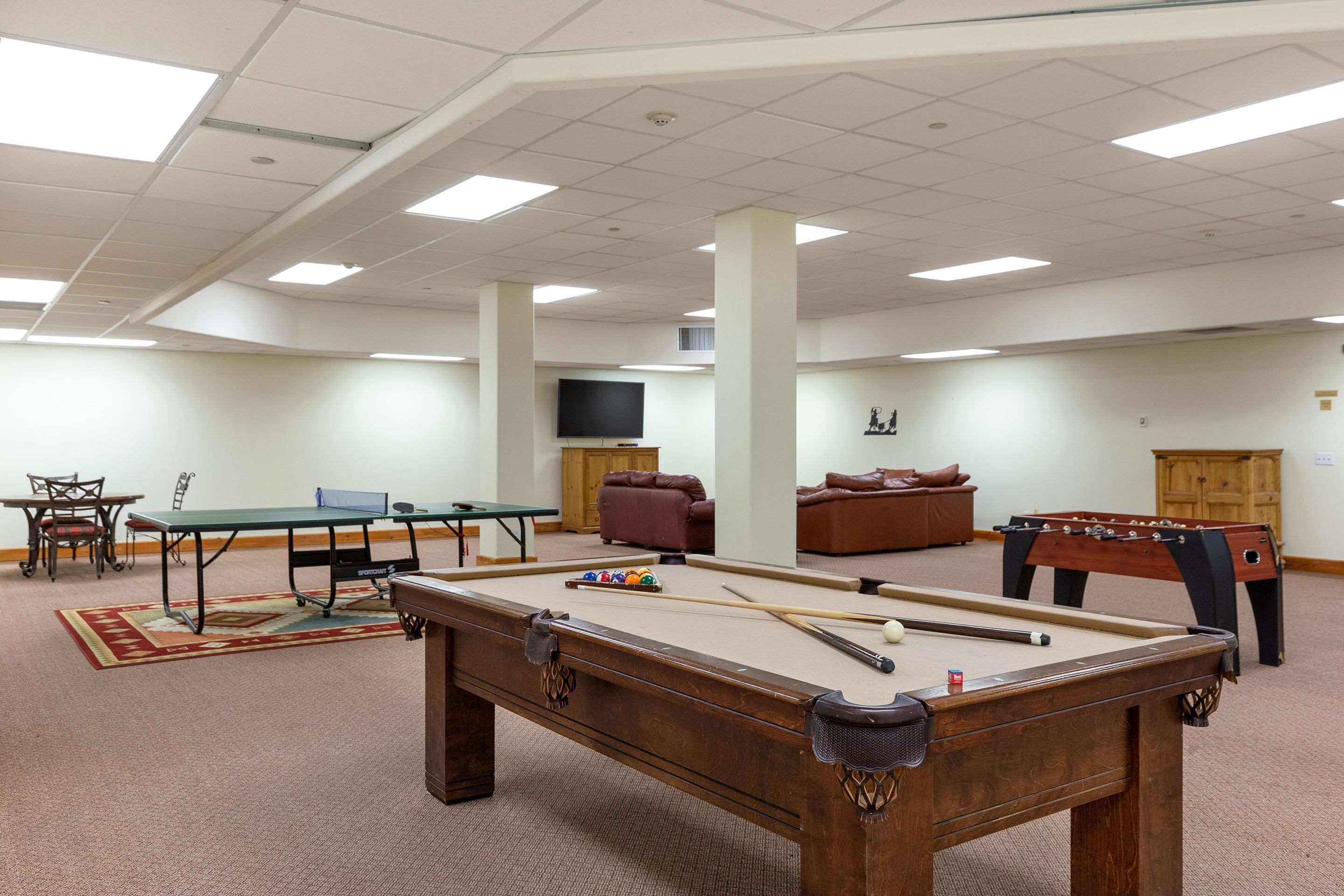 A pool table with billiard balls, a foosball table, a ping pong table, and a television in a room with beige walls and carpet.