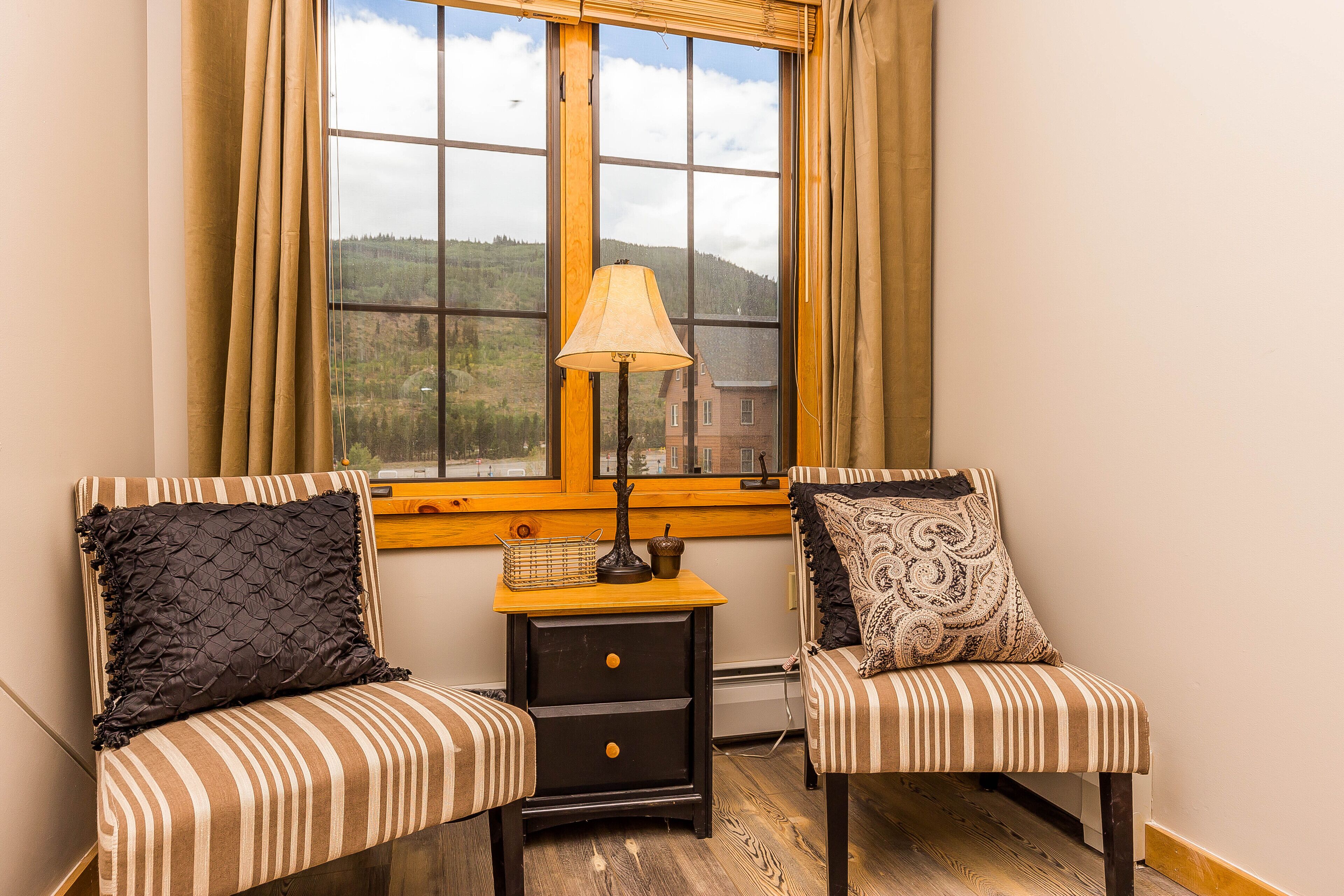 Two striped chairs with decorative pillows, a small wooden nightstand with a lamp, and a window with a view of a forested landscape.